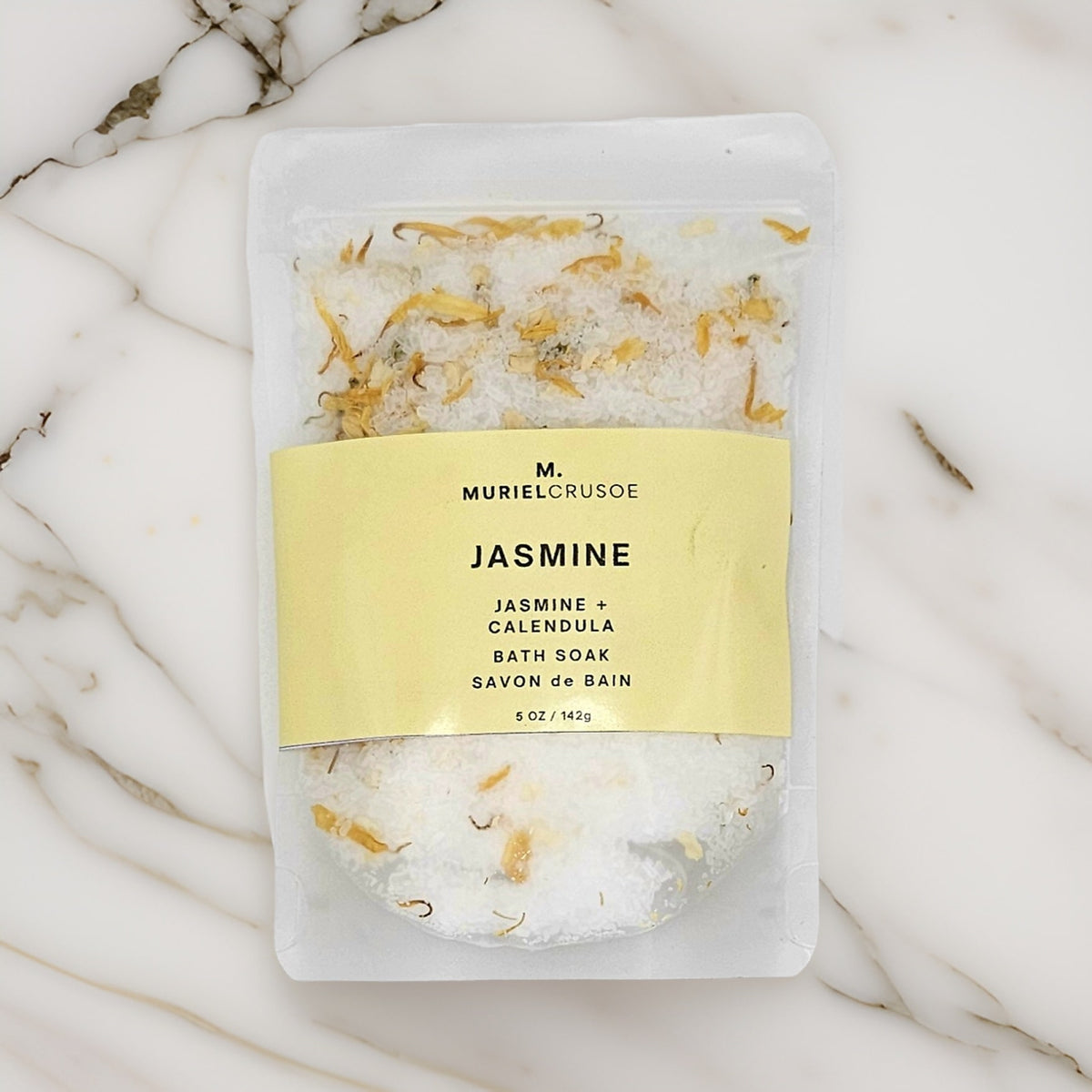 A pouch of Jasmine + Calendula bath soak with visible jasmine flowers and calendula petals, placed on a marble surface.