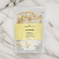 A pouch of Jasmine + Calendula bath soak with visible jasmine flowers and calendula petals, placed on a marble surface.