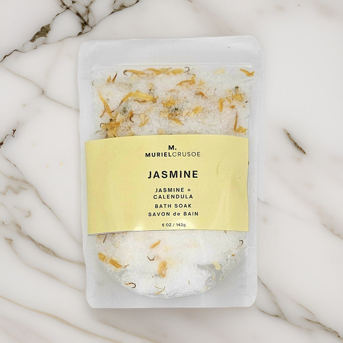 A pouch of Jasmine + Calendula bath soak with visible jasmine flowers and calendula petals, placed on a marble surface.