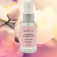 A spray bottle of JASMINE SUGARCANE PERFUME MIST with a pink label, placed against a blurred floral background.
