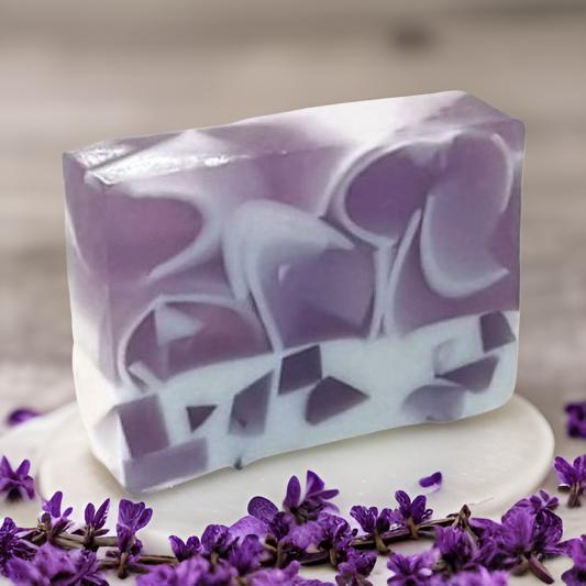 A bar of lavender vanilla soap with shea butter, displayed with scattered purple flowers around it.
